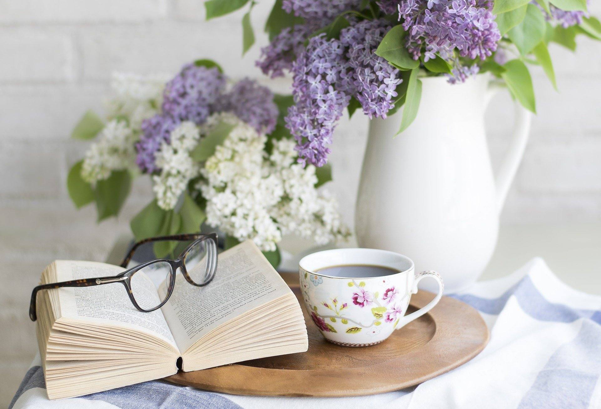 Auf einem hölzernen Teller liegt ein Buch, eine Brille, eine Tasse Kaffe. Im Hintergrund ist ein Blumenstrauß und eine Kanne Kaffee zu sehen.