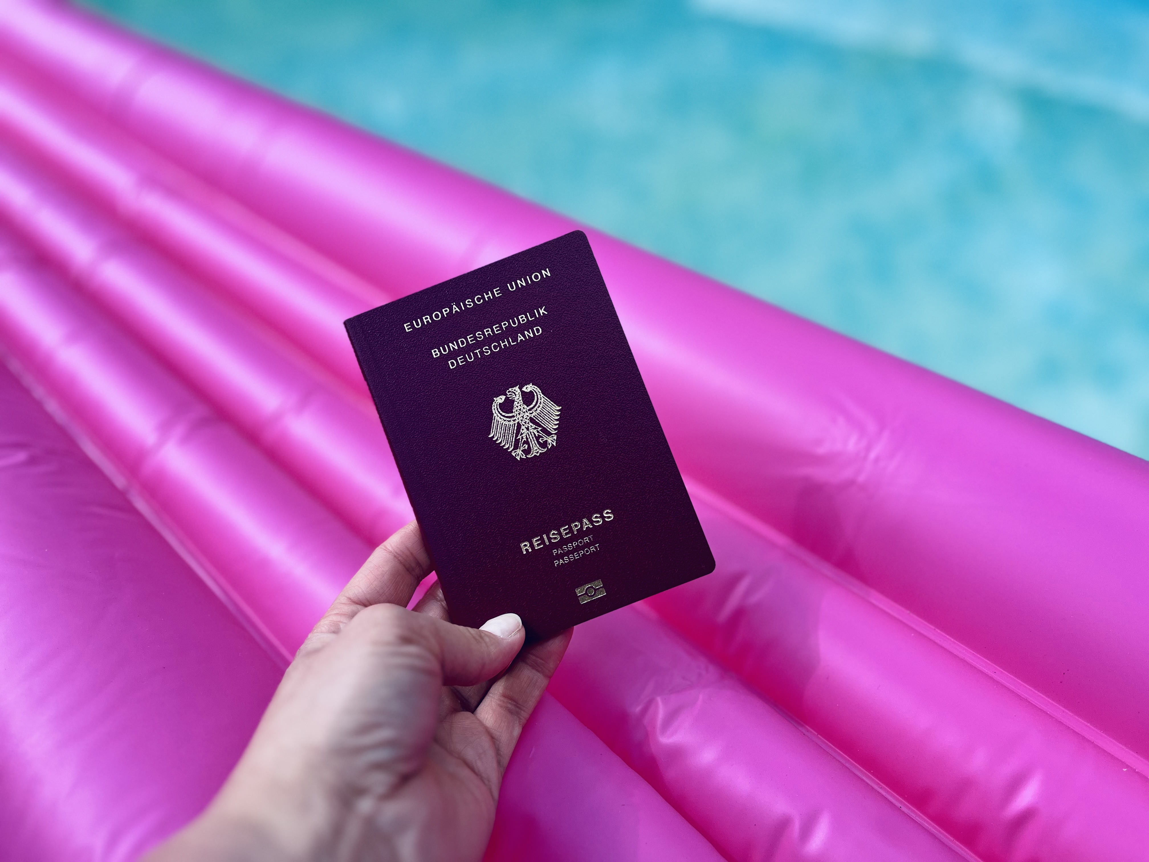 Hand hält Reisepass, im Hintergrund Luftmatratze im Pool