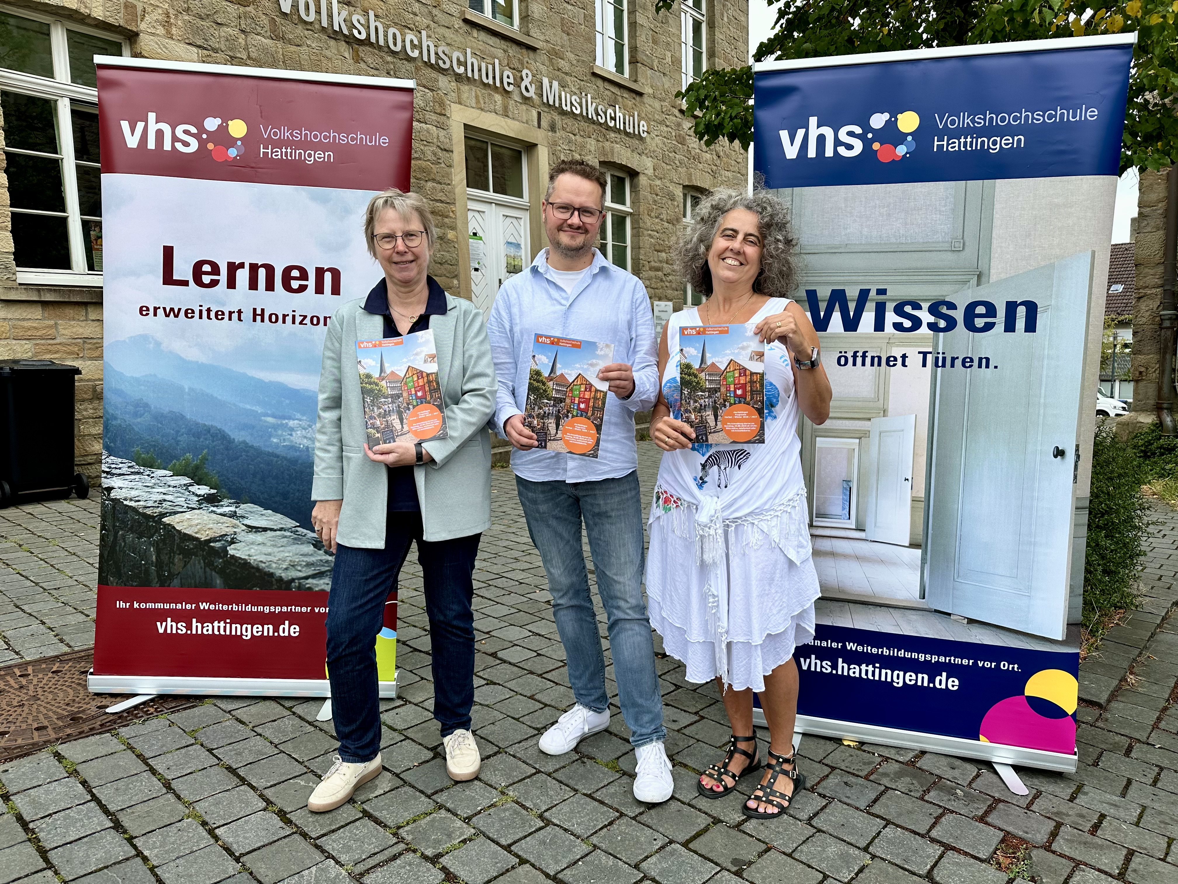 Drei Personen stehen vor zwei vhs Bannern und halten das Programmheft in den Händen. Im Hintergrund ist das Gebäude der vhs zu sehen.