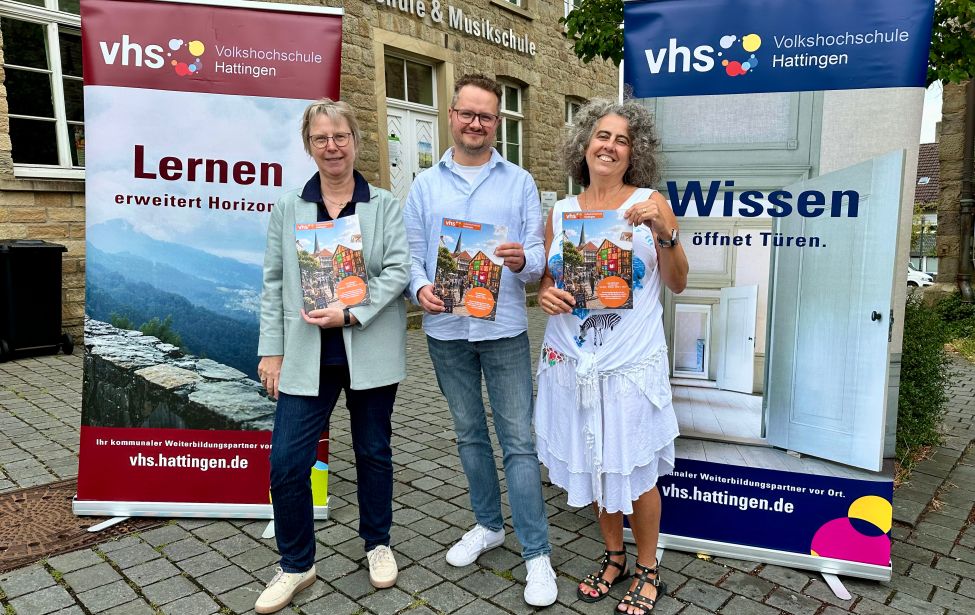 Drei Personen stehen vor zwei vhs Bannern und halten das Programmheft in den Händen. Im Hintergrund ist das Gebäude der vhs zu sehen.
